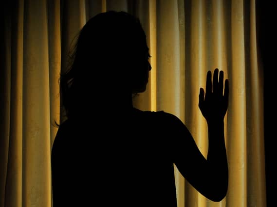 The silhouette of a woman standing in front of closed blinds, with one hand touching the blinds